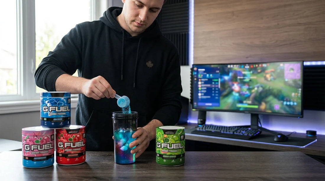 A man in a black hoodie scoops blue G Fuel powder into a shaker bottle at a gaming desk with a monitor and tubs of energy…